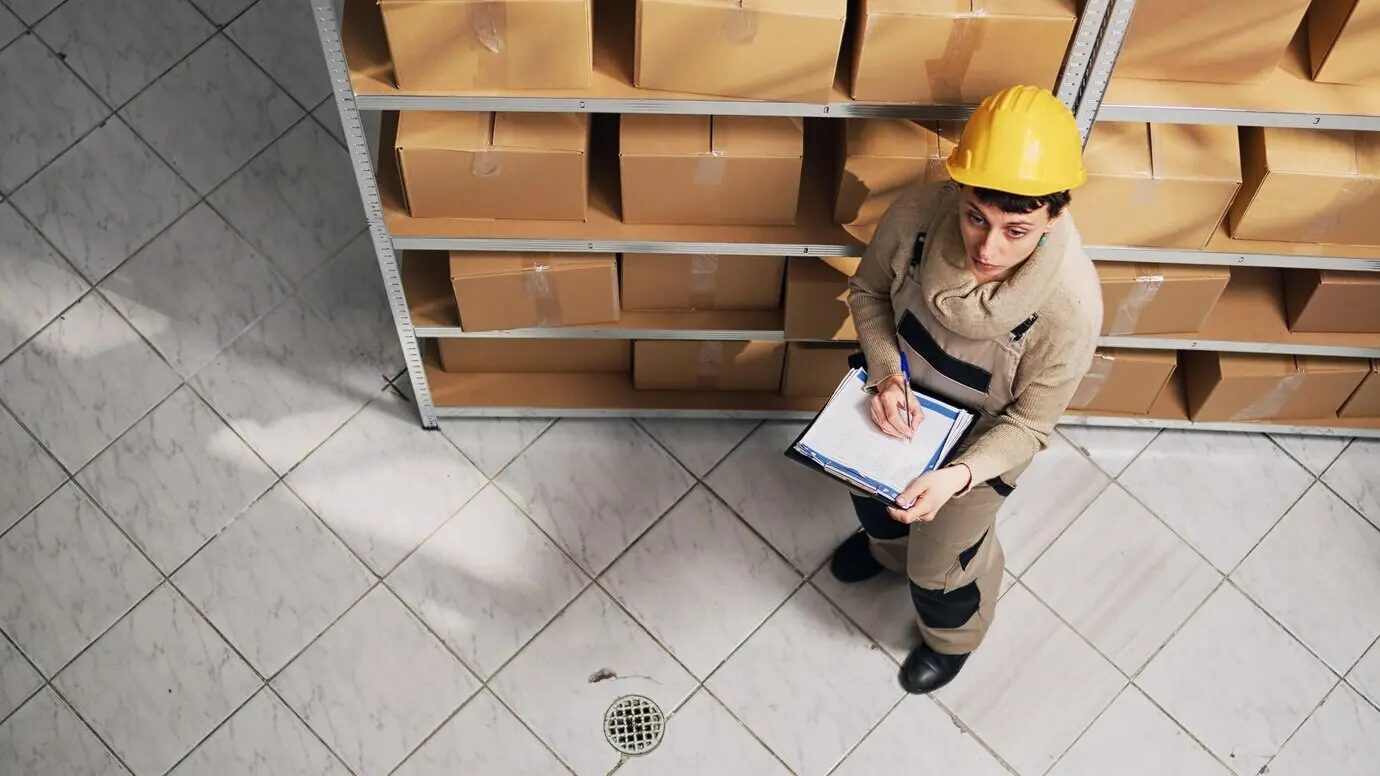 Менеджер склада изучает список товаров на полках, смотрит на картонные упаковки, чтобы спланировать отправку. Руководитель в комбинезоне работает в складском помещении с товарами и грузом, занимаясь складской логистикой.